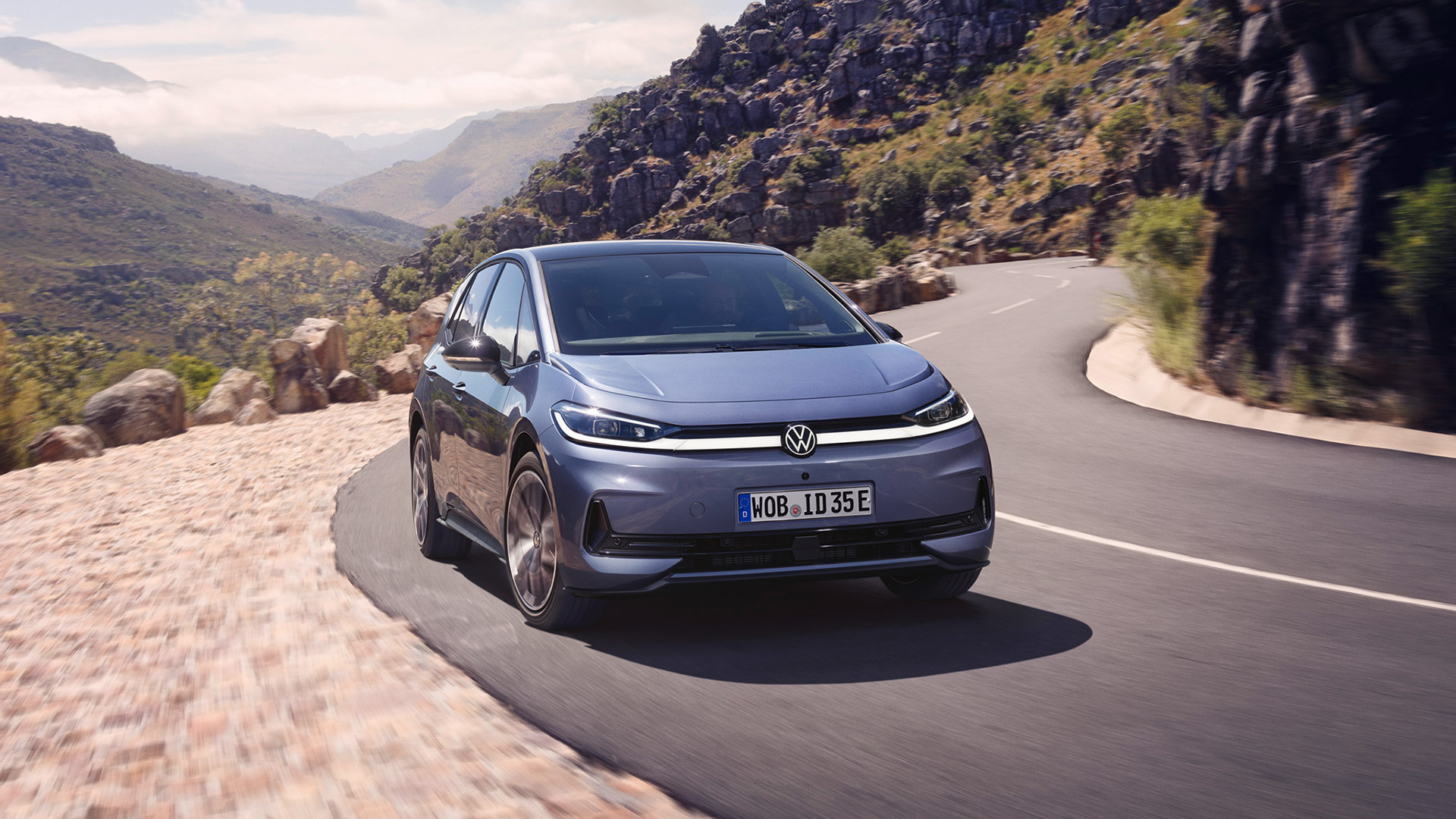 Blauer ID. 3 Neo f&auml;hrt auf einer kurvigen Landstra&szlig;e. Im Hintergrund Berglandschaft. Vorderansicht. 